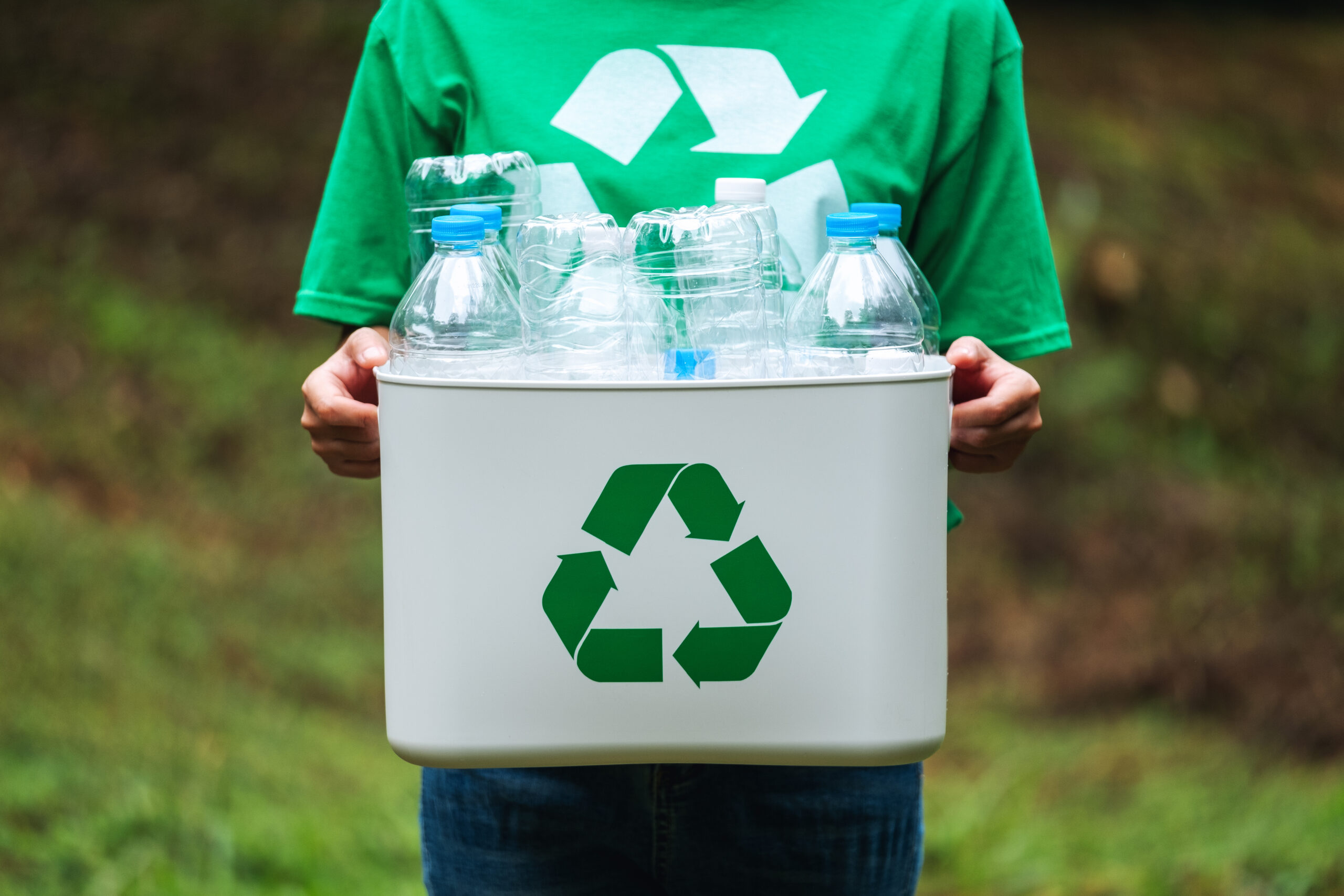 A woman in green holding a recycle bin with plastic bottles in the outdoors.