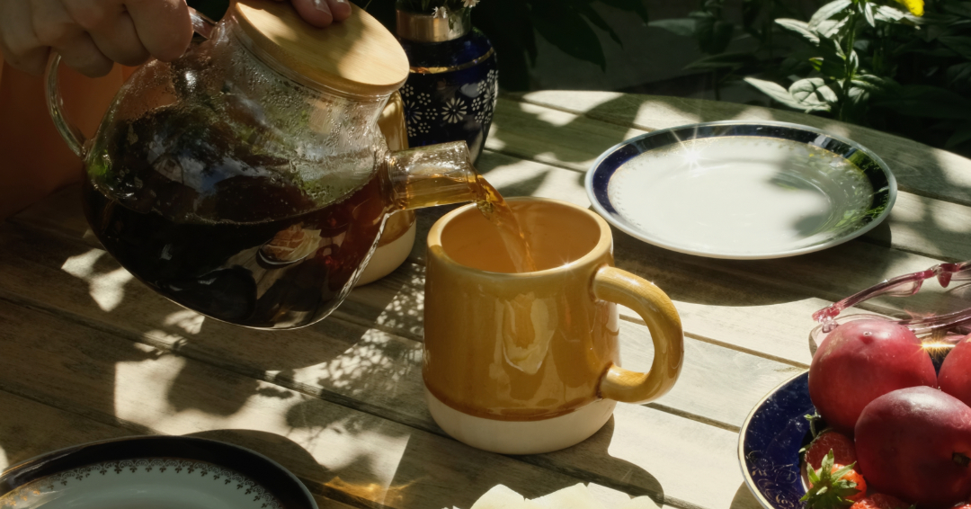 Someone pouring tea into a ceramic mug on a wooden table. The tea party is outside and the sun is shining.