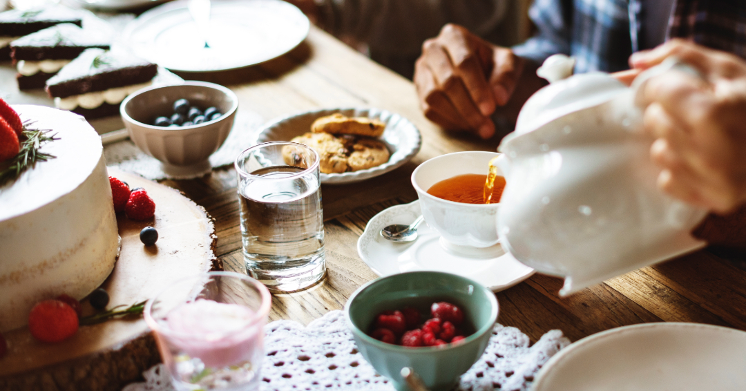 People gathered around a tea party table, with berries, cookies, and a cake. Someone is pouring tea.