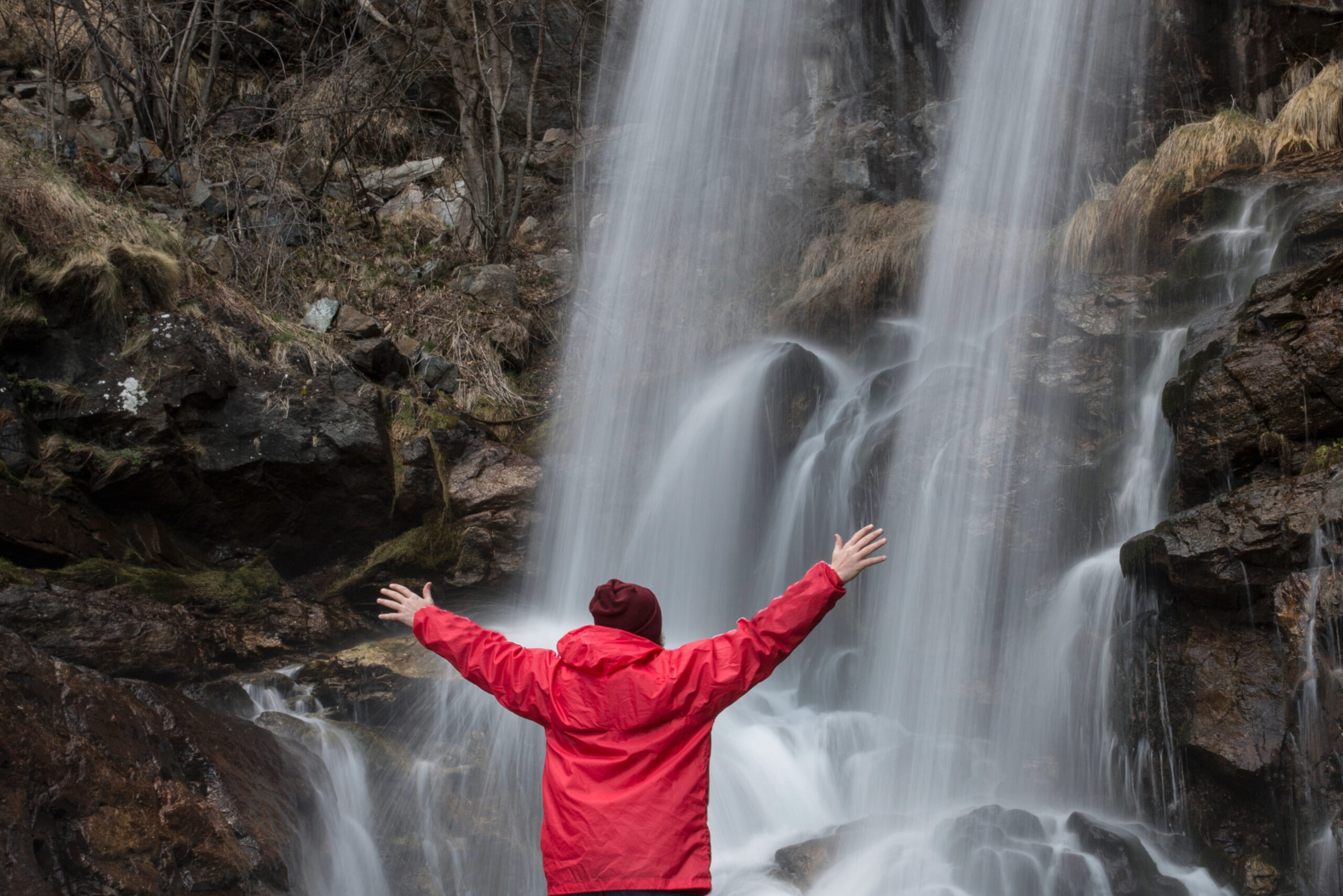 Person in a red coat, arms stretched out wide, facing a cascading waterfall.