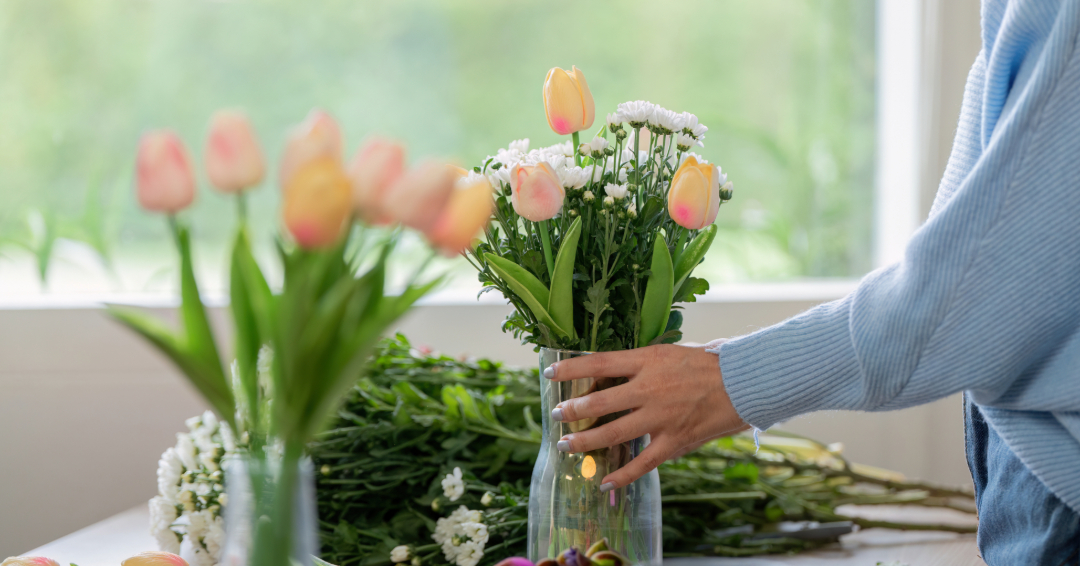 A woman, wearing a blue sweater, touching a vase of light pink tulips. 