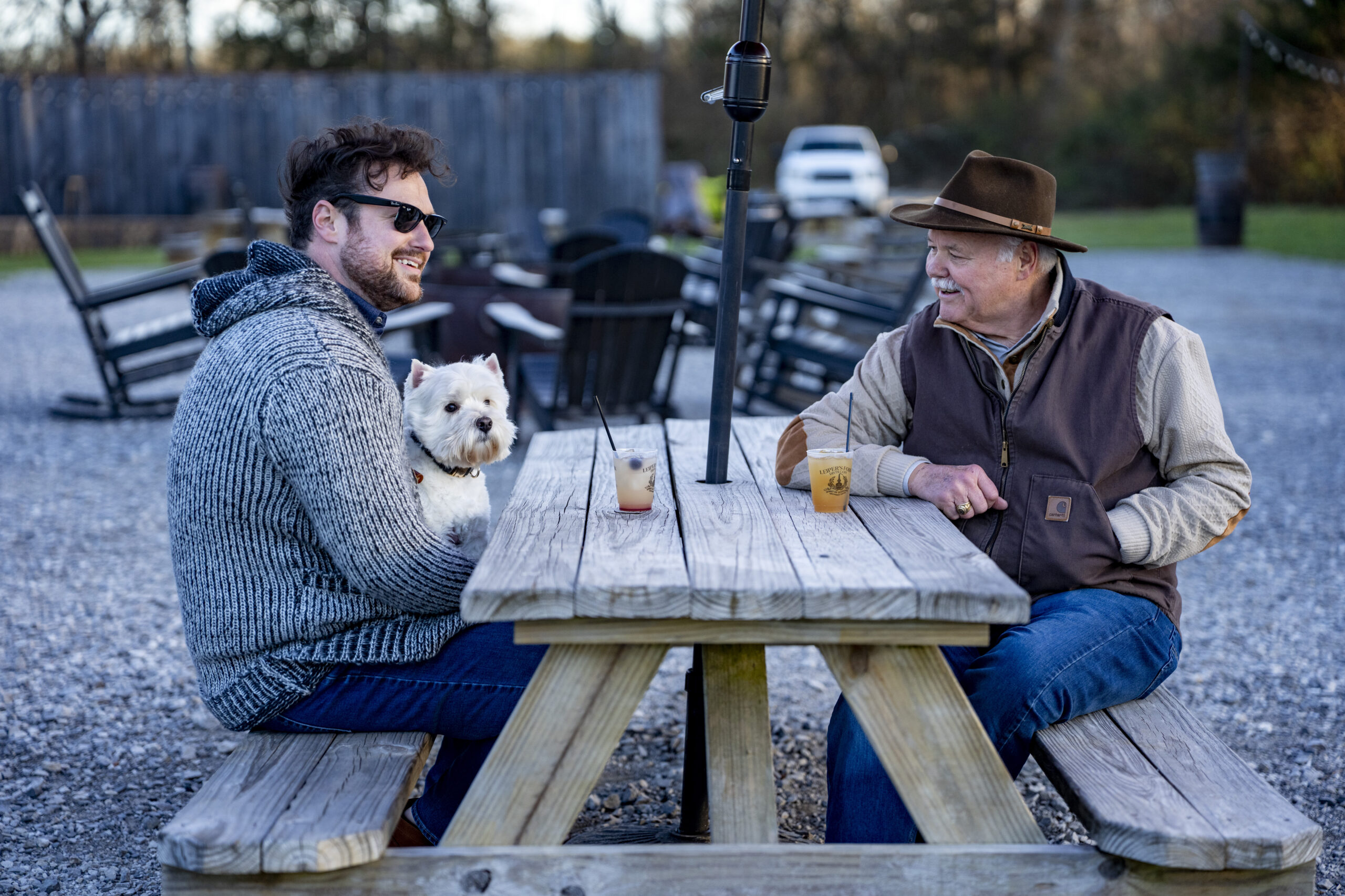 Whiskey & Wags Two Rivers Mansion, two men sitting at a picnic table with a small white dog during an outdoor whiskey tasting event in Nashville.
