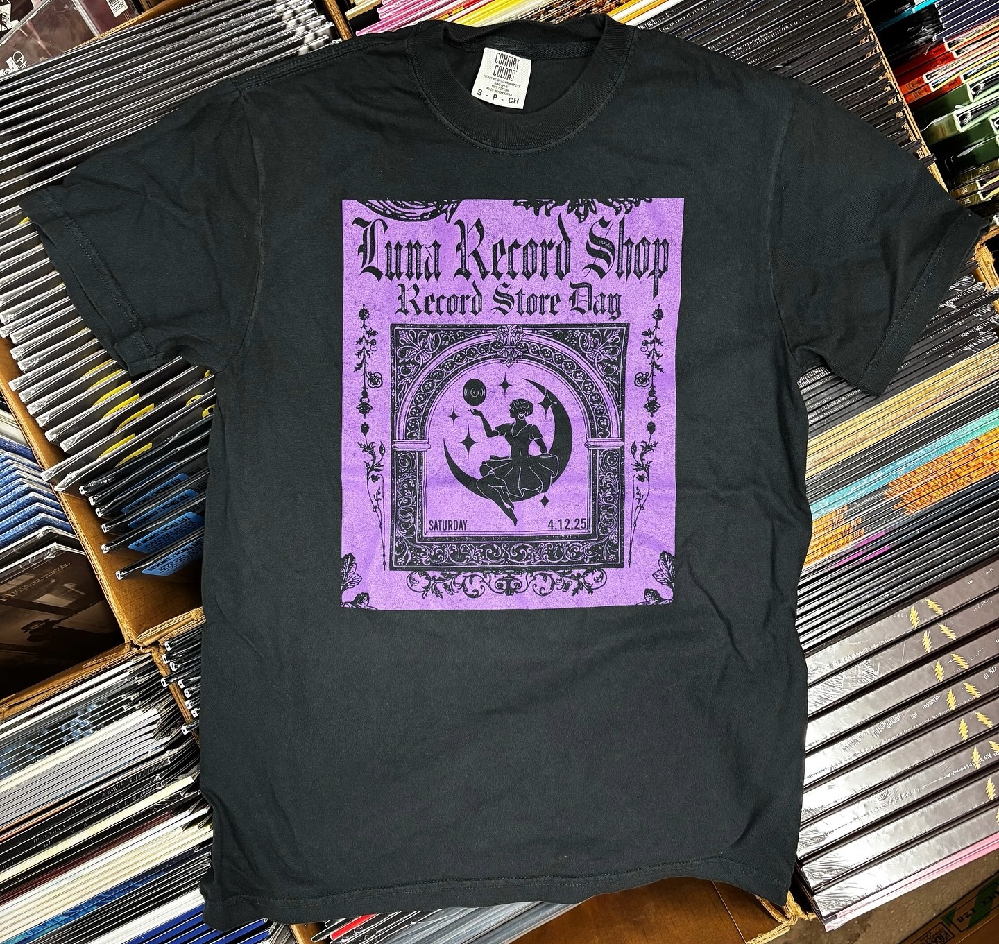 Black Luna Record Shop Record Store Day t-shirt with purple graphic displayed on stacks of vinyl records at The Factory at Franklin
