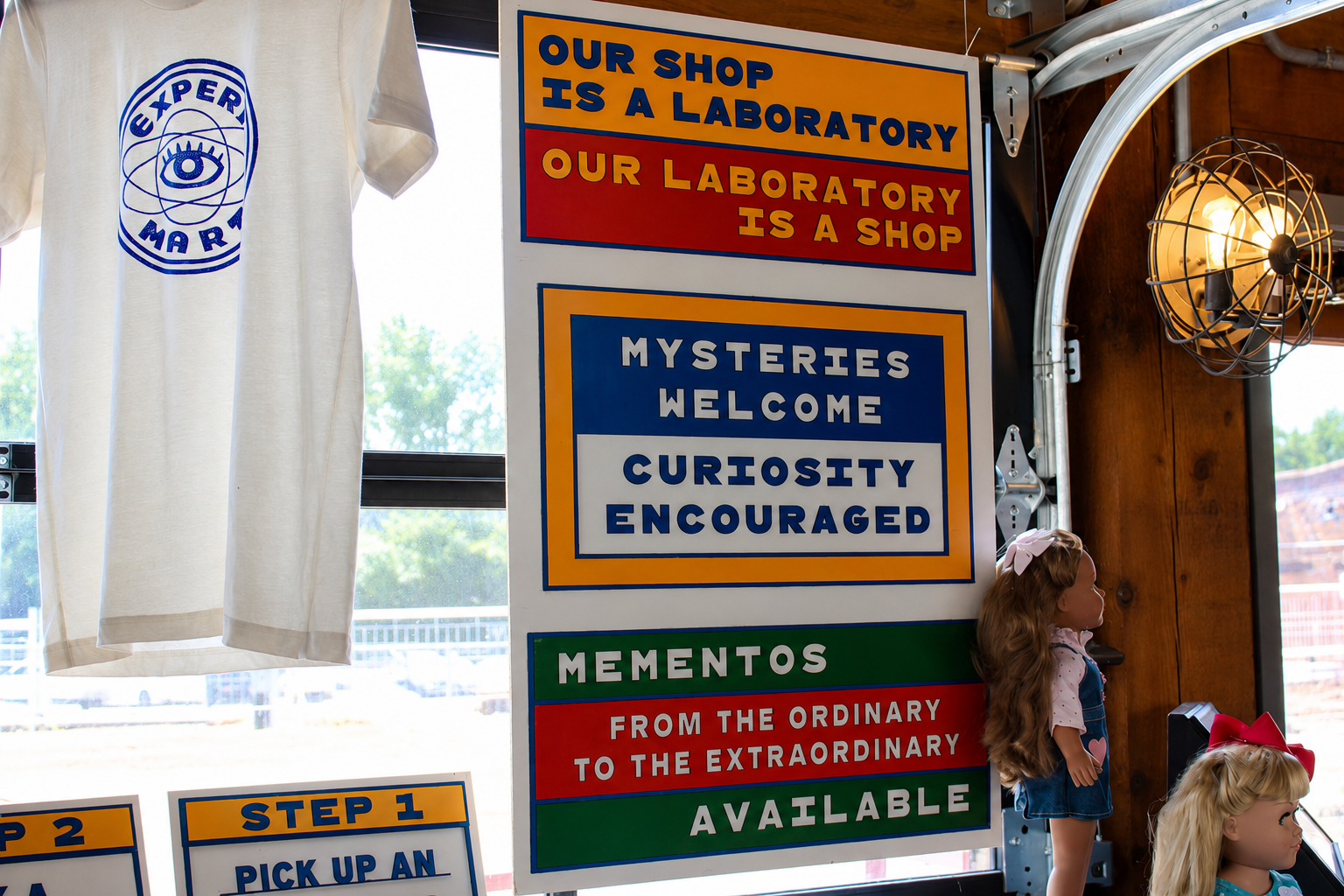 Experimart T-shirt and signs reading “Our Shop Is a Laboratory,” “Mysteries Welcome,” and “Curiosity Encouraged”