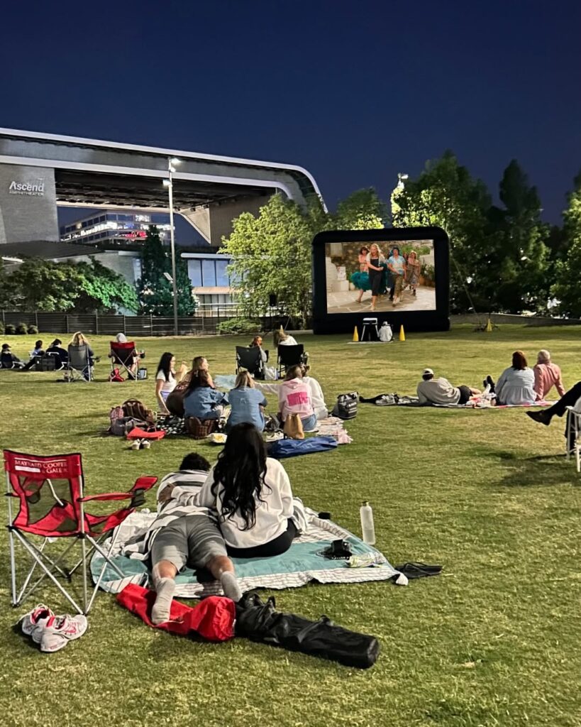 People watching an outdoor movie on the lawn at Riverfront Park in downtown Nashville, Tennessee, with Ascend Amphitheater in the background.