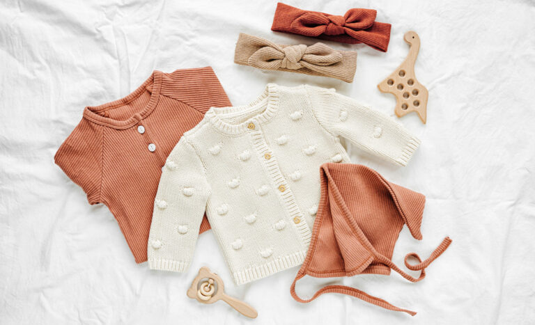 Set of baby rompers, hat, hairband and knitted jumper on white bed. Fashion baby clothes and accessories. Flat lay, top view