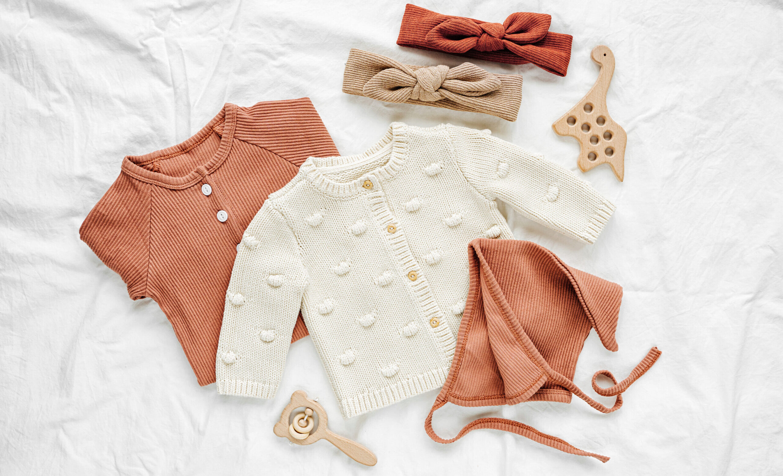 Set of baby rompers, hat, hairband and knitted jumper on white bed. Fashion baby clothes and accessories. Flat lay, top view