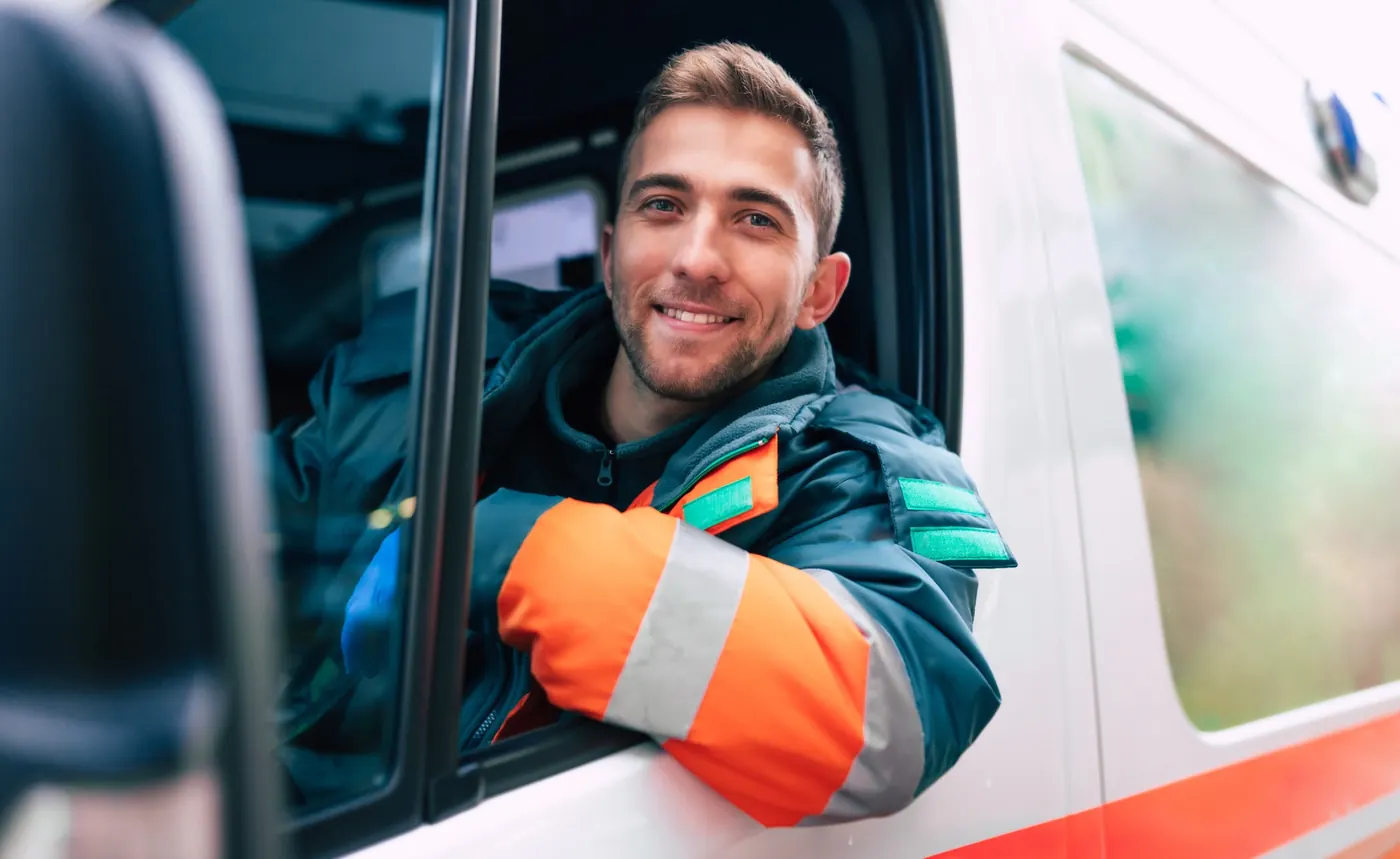 Smiling emergency medical responder wearing a reflective uniform while seated in the driver's seat of an ambulance.