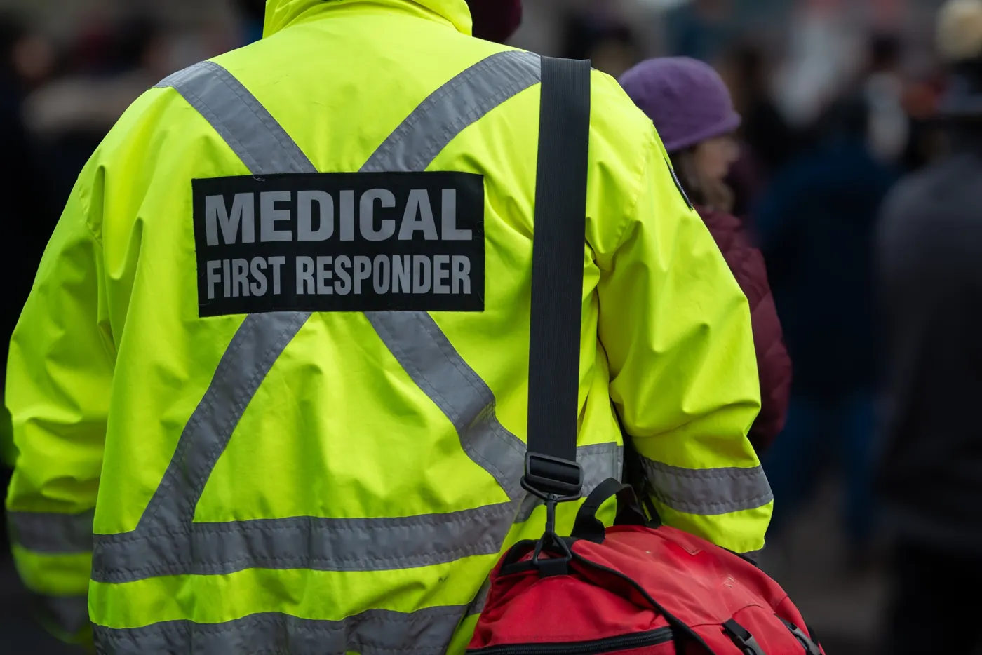 Medical first responder wearing a high-visibility yellow jacket with reflective stripes and carrying a red emergency medical bag.